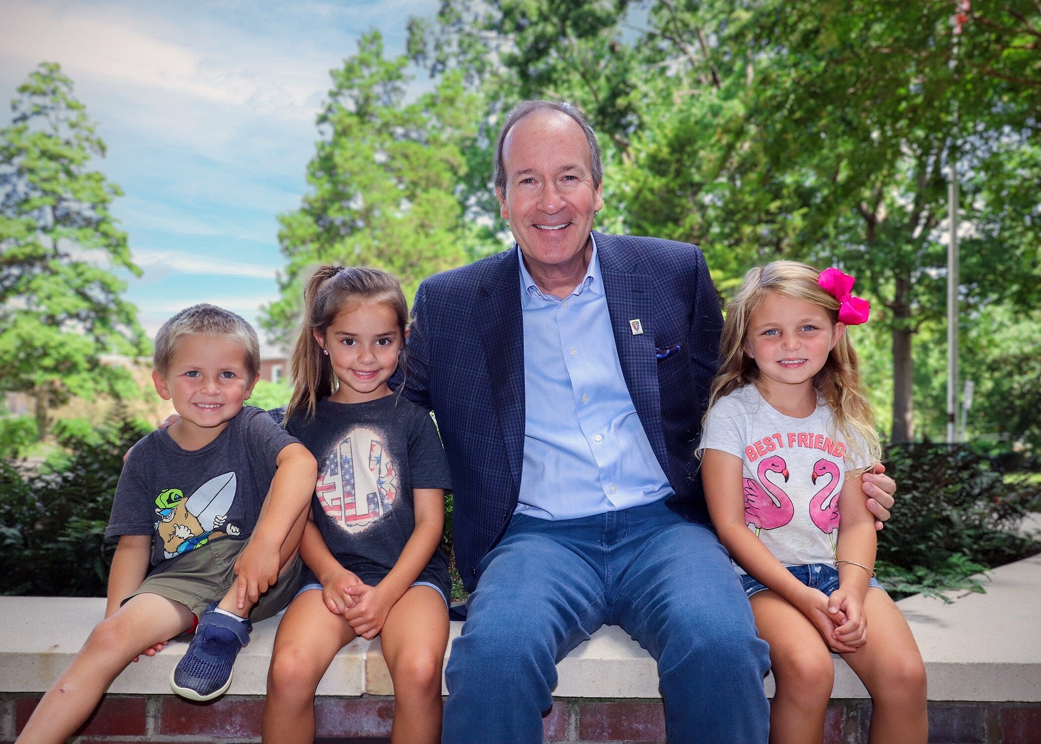 A man in a suit jacket and jeans sitting on a stone ledge outdoors, flanked by three smiling young children.